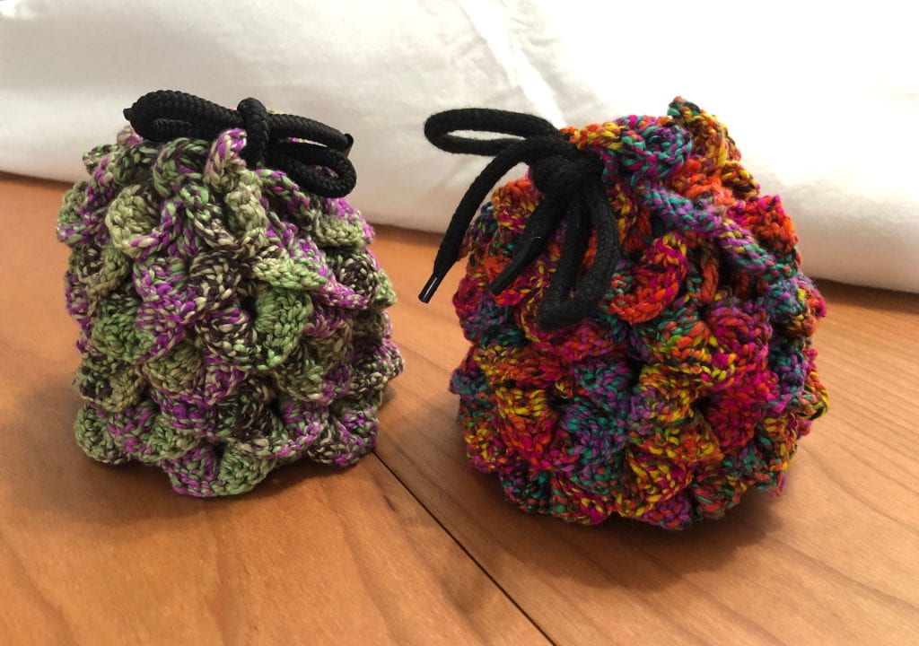 Two crochet drawstring bags. One mottled green/purple, one variegated rainbow. The bags have overlapping "scales" that make them look like a dragon egg or a pinecone.