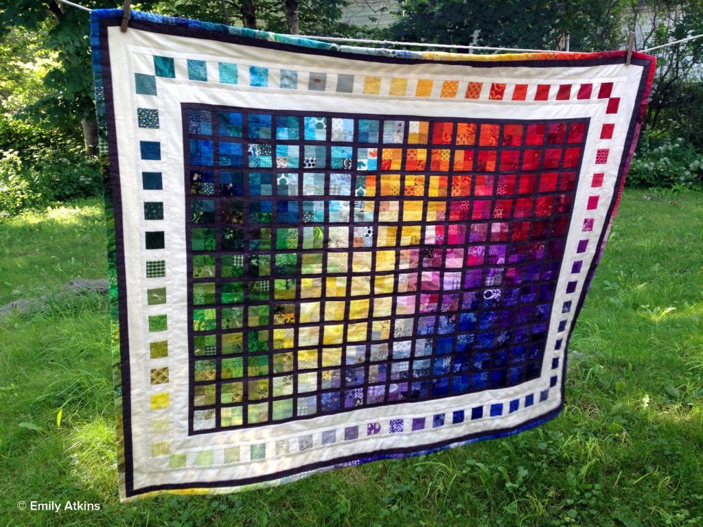 A rectangular quilt hung outside on a clothesline. The vibrant rainbow colors swirl and bleed together and are set within a traditional windowpane pattern in charcoal and are bordered by white inset with rainbow squares.