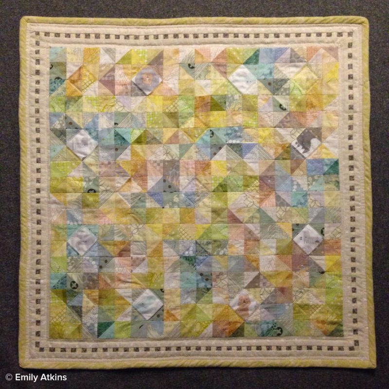 A square pastel quilt on a gray background. The random use of color created a scattered mosaic appearance with the traditional pinwheel block pattern, the body of the quilt is bordered by white with alternating light and dark squares running along the center of the border.