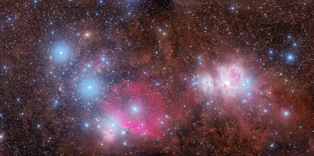 Millions of stars shine in the night sky with white, warm yellow and blue colors. Dust nebulosity of brown and pink is scattered around. Three white-and-blue-lit stars in a line seem bigger than the rest.