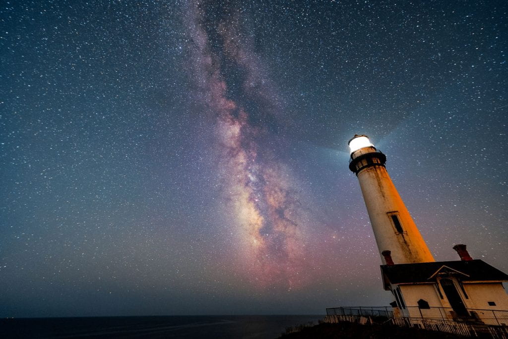 The light of a white-walled lighthouse shines bright under a night sky were millions of stars are visible, along with the pinkish layout of the milky way.