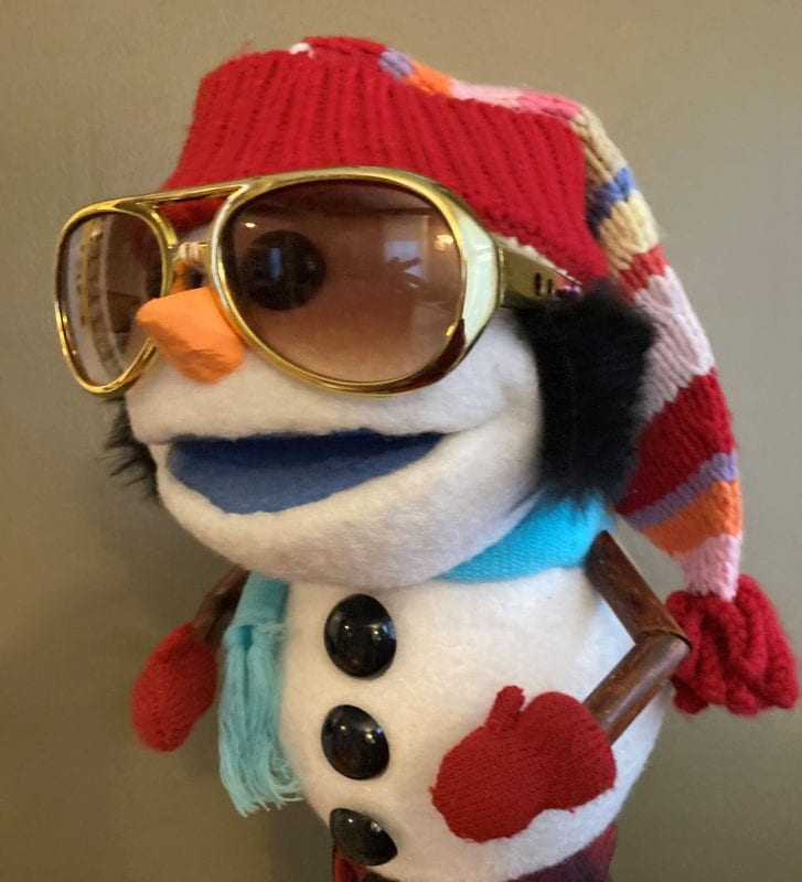 A snowman puppet that is dressed as an Elvis impersonator. Long stocking cap hat, blue scarf, and Elvis glasses on a round snowman with a tiny carrot nose.