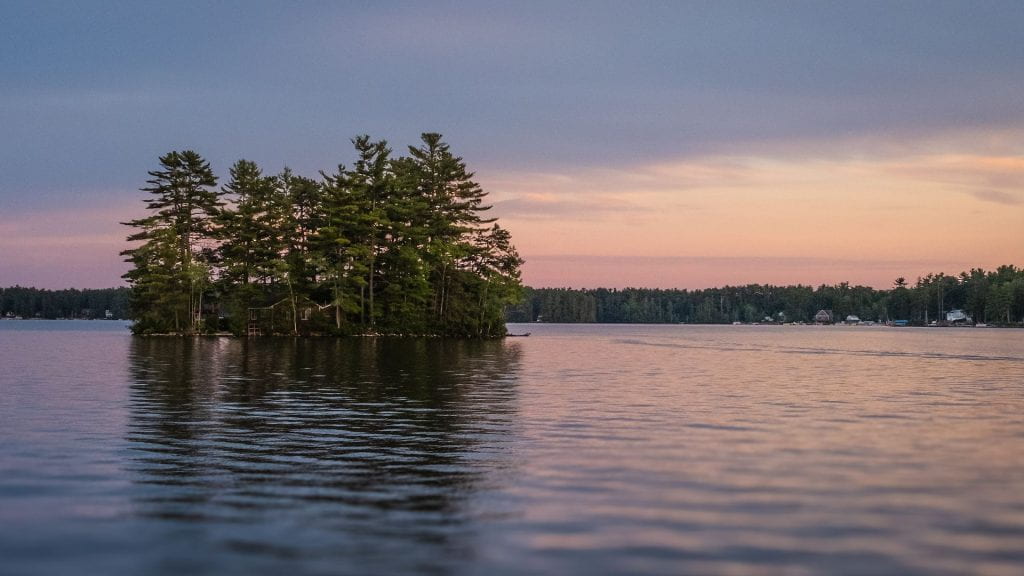 A small island with tall pine trees and a cabin, in a lake with a purple sky.