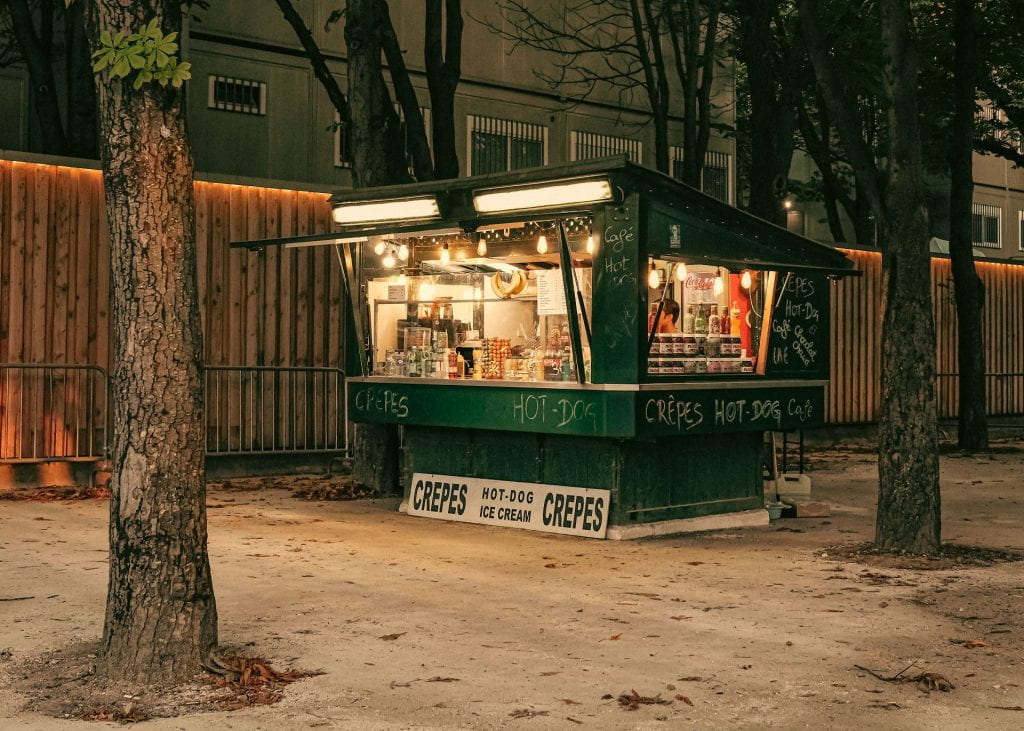A dark green crepe & hot dog stand at dusk, with its lights on.