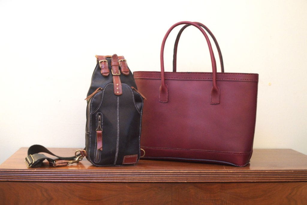 Two leather bags rest atop a wooden table. One is a deep magenta tote with detail stitching in the same color at top and bottom; the other is a black shoulder or camera bag with white detail stitching, brown buckles, strap, and zipper pulls.
