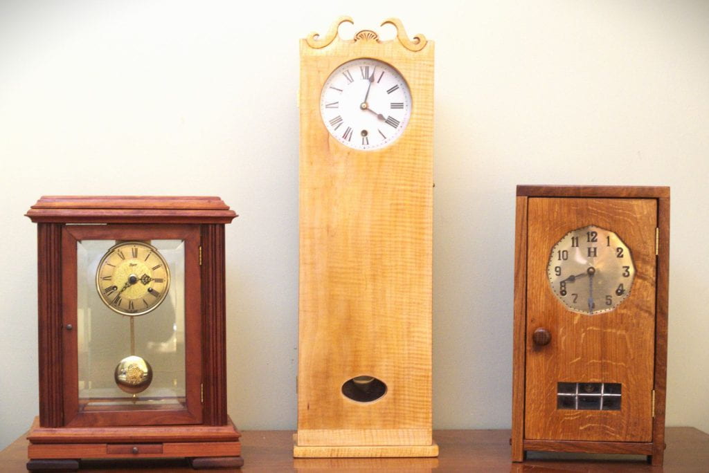 Three analog clocks. The leftmost has glass panes in the center, a gold pendulum, and red wood. The center is a tall clock of pale figured maple, with curlicues at the top and an internal pendulum just visible in an oval opening. The rightmost is made from warm brown oak, rectangular, with a knob for opening the center panel and a grating at the bottom of the panel.