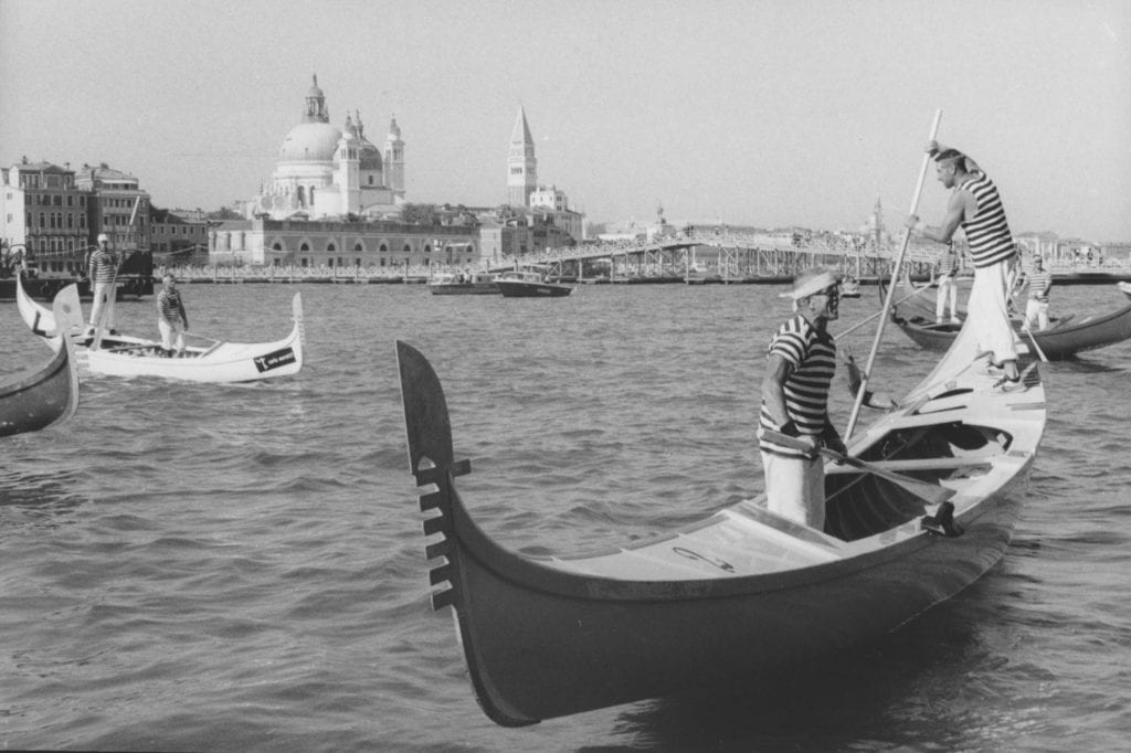 Black and white photograph of three gondolas on the water and buildings of Venice visible in the background. Two gondoliers with t-shirts of black and white stripes are on each boat.
