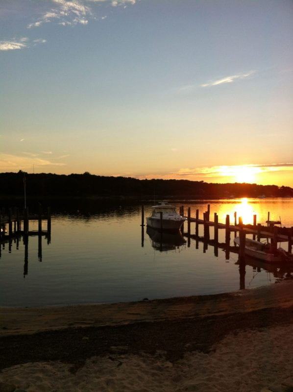 The sun is setting in the distance, overlooking a calm lake with a sandy beach and two wooden piers. Two small boats are visible docked in one of the piers. 