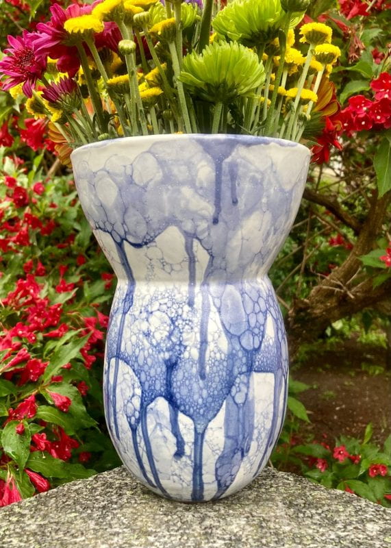 A curved, white vase with blue bubble and drip designs, holding white and pink flowers in a garden.