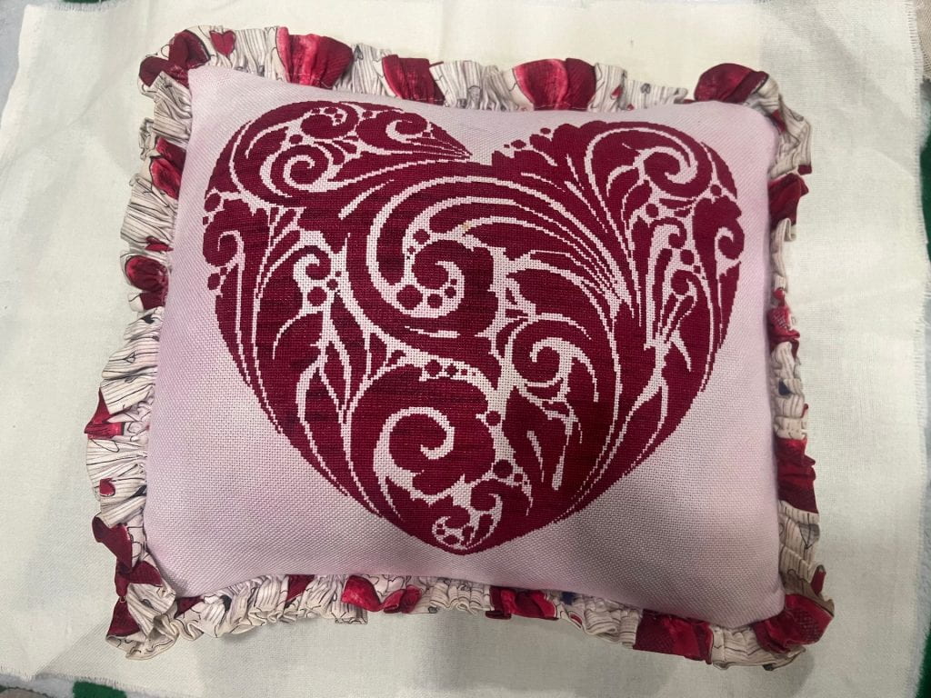A heart cross-stitched on light pink fabric, stitched in a deep red thread. It is finished as a pillow with a trim around the border, in a fabric with hearts.