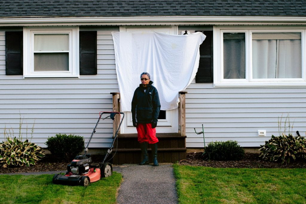 A black woman with a short haircut is posing in front of a house entrance and standing near freshly mowed garden lawn and a few garden shrubs. She is wearing sunglasses, black rubber boots and waterproof attire with a black jacket and red pants. The entrance behind her is covered by a white sheet and the house's white wooden panel walls and black tiled roof are also visible.