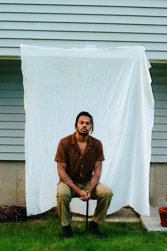 A black man is posing seated on a stool for a portrait picture. His hands are crossed on his lap. He is wearing a brown short-sleeved shirt, khaki pants, and black shoes. There are a number of tattoos on his arms. The white wall wooden panels behind him are covered by a white sheet.