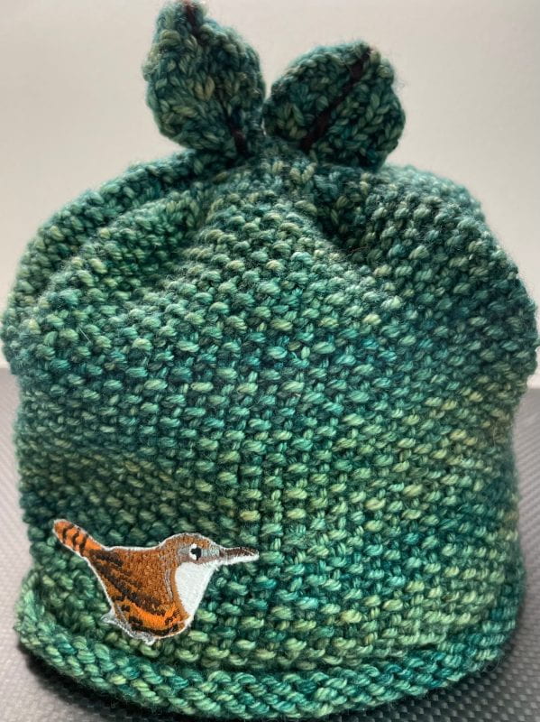 A knitted forest green winter hat with a rolled rim, leaves at the top, and a wren bird patch at the base.