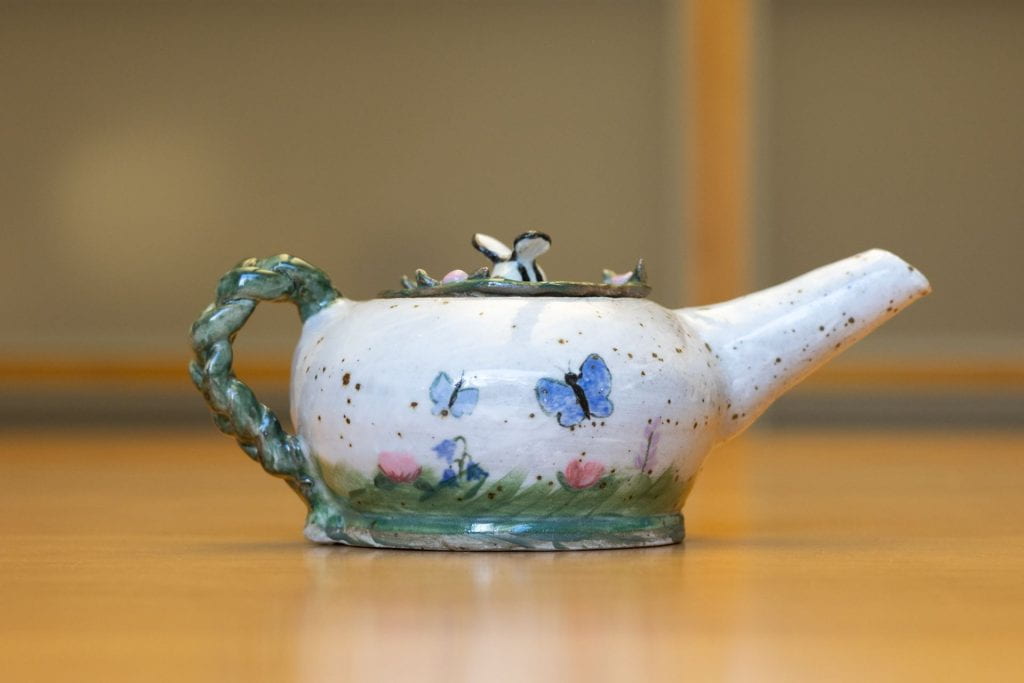 A ceramic teapot patterned with flowers, bees, and butterflies, with a green vine handle and a bee on the lid.