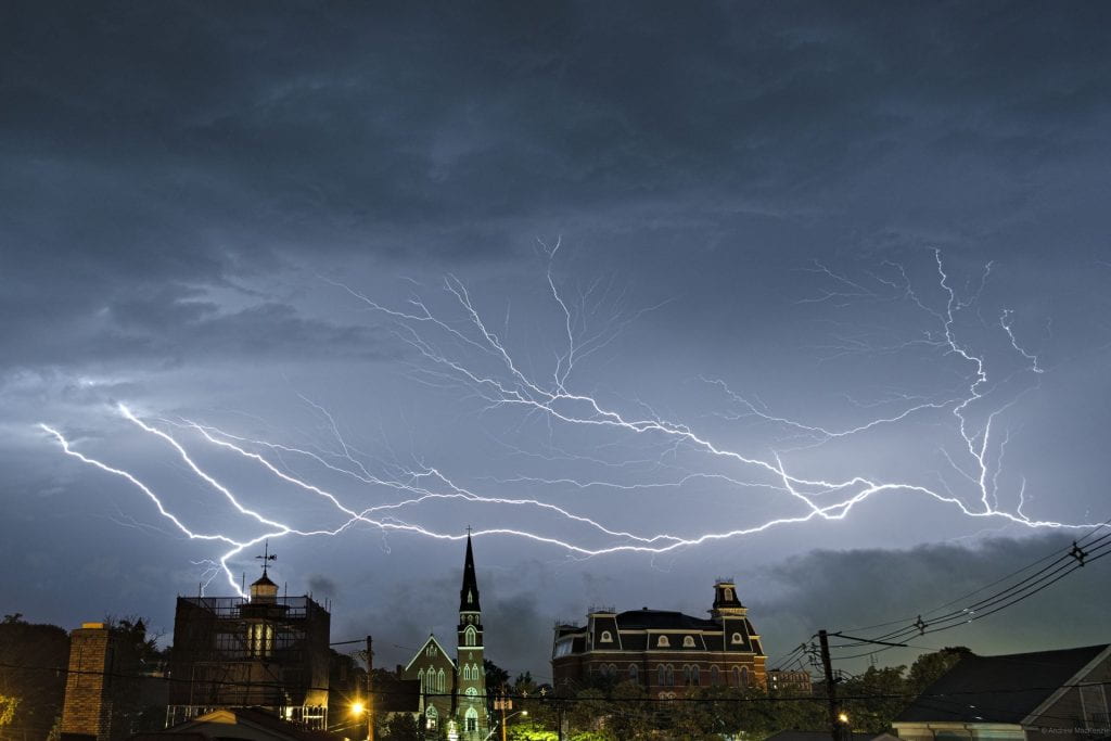 A brilliant lightning bolt scatters across the sky above city hall and a church. The lightning has many forks with small tendrils which illuminate the cloudy sky.