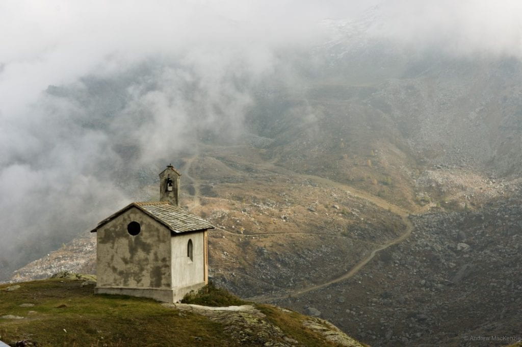 A photograph looking past a chapel into a valley in the alps. A winding road is seen in the valley floor beyond.