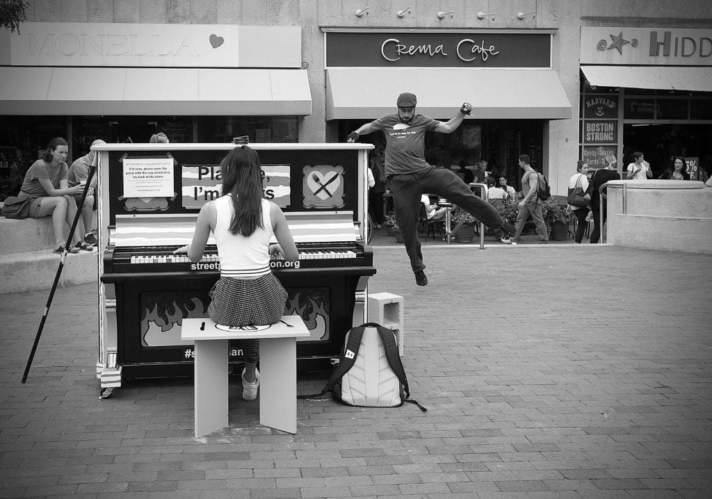 A woman plays an outdoor piano while a man dances and is caught in mid-air in busy Brattle Square, Cambridge.