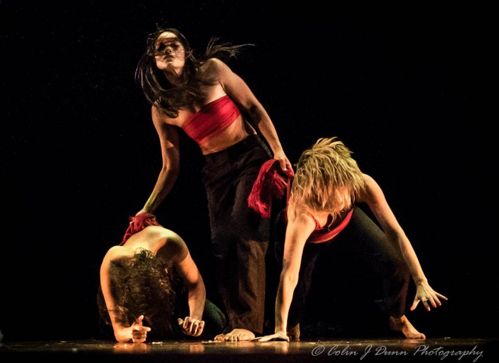 Three dancers with red bands around their chests and black pants, caught in the middle of a twisting, bending dance.