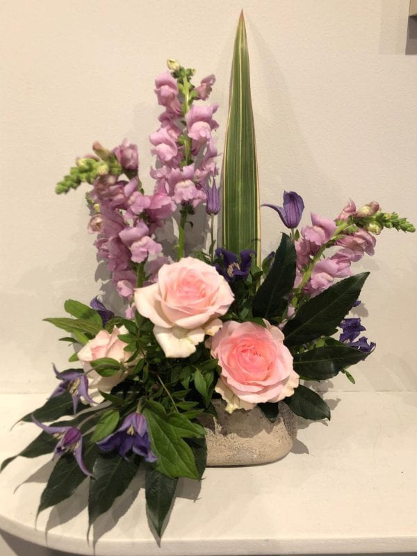 Floral arrangement with lush pink roses, pale lavender snapdragons, deep purple clematis, and striking dark green leaves.