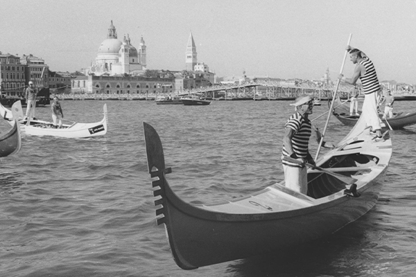 Black and white photograph of three gondolas on the water and buildings of Venice visible in the background. Two gondoliers with t-shirts of black and white stripes are on each boat.   