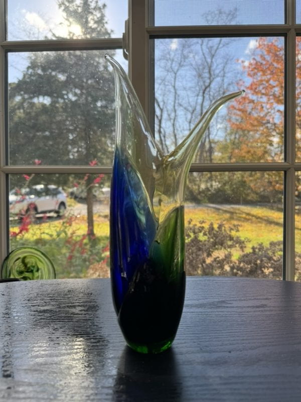 This glass piece works by dipping a clear vessel into first a pot pot of molten glass and then into a green pot. I then pull two sides. The blue tends to pull further as it is considered a softer color. The green compliments the with its own blue components.