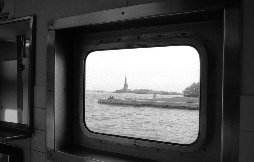 The Statue of Liberty is framed by the metal window frame inside the bathroom of the Ellis Island Ferry. Image is in black and white.