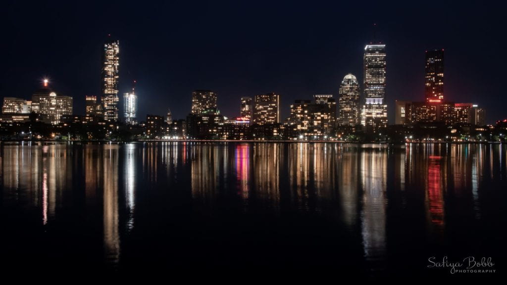 A view of the Boston skyline taken at night. The sky is nearly black. The lights from the buildings are reflected in the Charles River below.