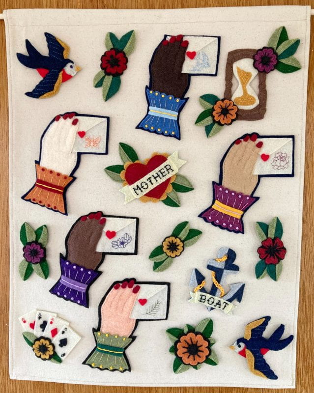 Jewel-toned patches arranged on a rectangular tan background hanging on the wall: two sparrows on opposite top and bottom corners; an hourglass; five hands in different skin tones holding envelopes sealed with a heart; a heart with ruffles, leaves, and a banner across the middle that says “Mother”; a two-toned anchor with a crochet chain and a banner that says “Boat”; a large orange flower with five heart-shaped petals and eight leaves around it; and similar smaller flowers with two leaves on either side, interspersed around the other designs.