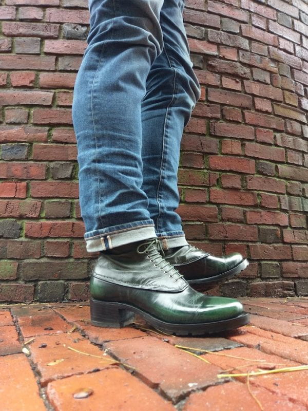 Close-up of the legs of a person wearing cuffing denim jeans and iridescent dark green leather boots, against a background of brick wall and pavement.