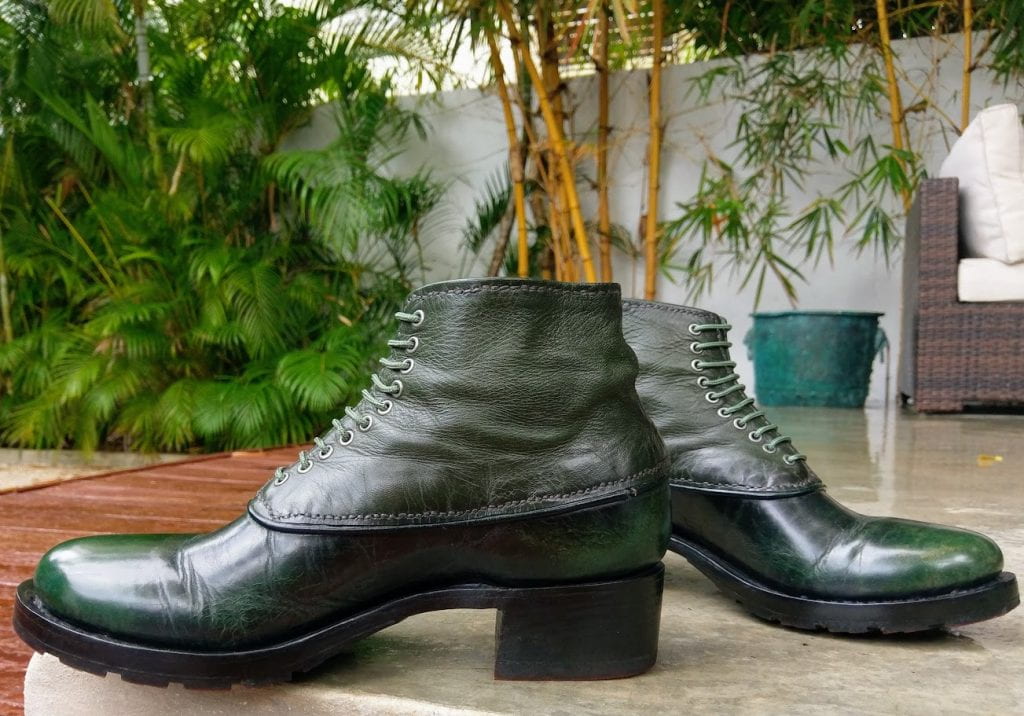 Iridescent dark green leather boots with olive-colored laces and darker heels and outsoles, against a background of bamboo plants.