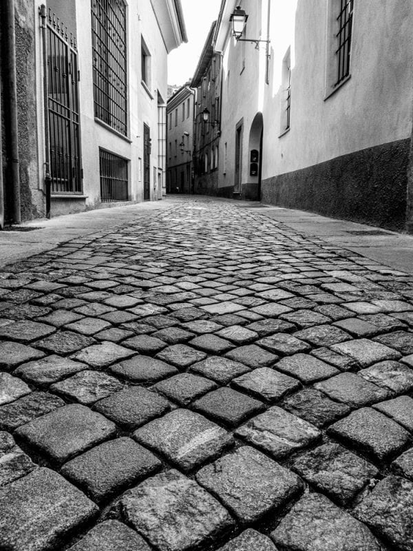 Black and white photo of cobblestones on a street in Italy taken from an angle that is close to the street with buildings in the background.