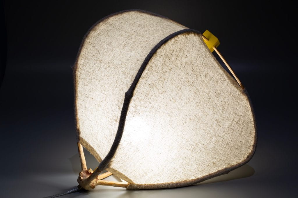 An illuminated lamp with irregular rounded sides formed from the curved branches of a young tree. The bent branches are covered with an open weave linen fabric and their tips are bent together and fitted into four sides of yellow child's building block.