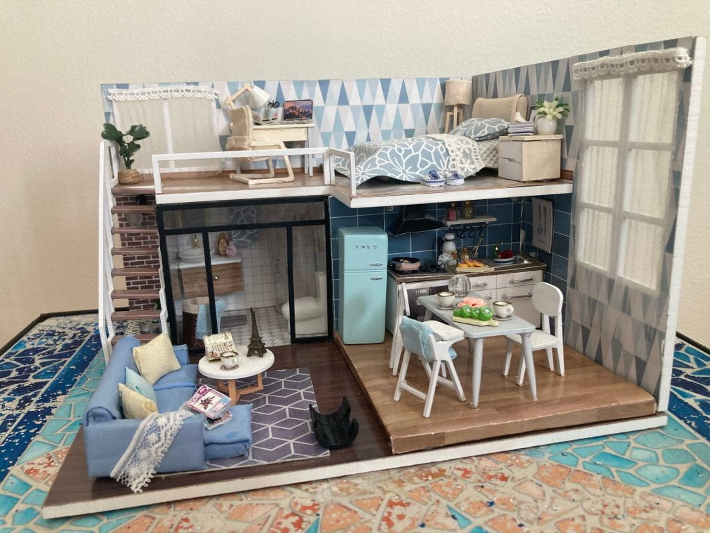 An cutaway view of a miniature two-story apartment. The apartment has a kitchen, living room, bathroom, and bedroom, furnished throughout in many shades of blue and white.