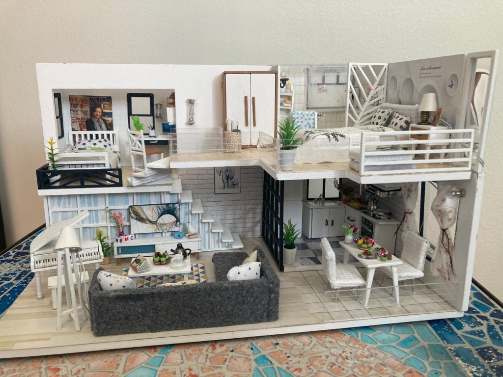 An inside view of a miniature three-story apartment, furnished mostly in white. Its rooms include a kitchen and living room on the first floor, a bedroom on the second floor, and a bathroom and bedroom on the third floor.