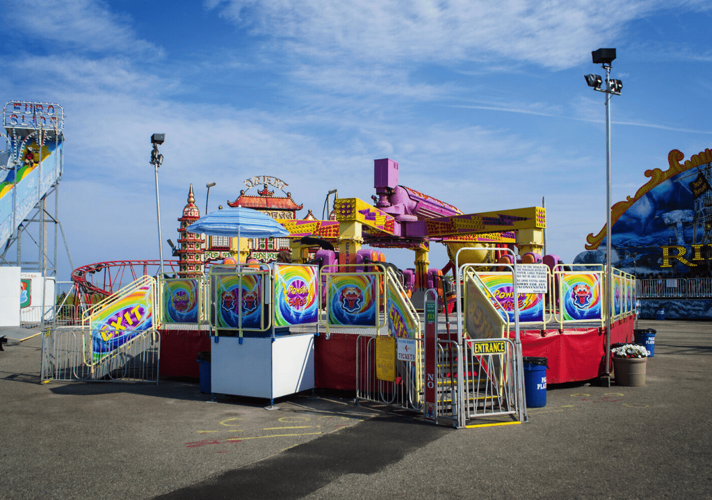 A brightly colored carnival ride, with a raised platform, sits on asphalt with other rides in the background.