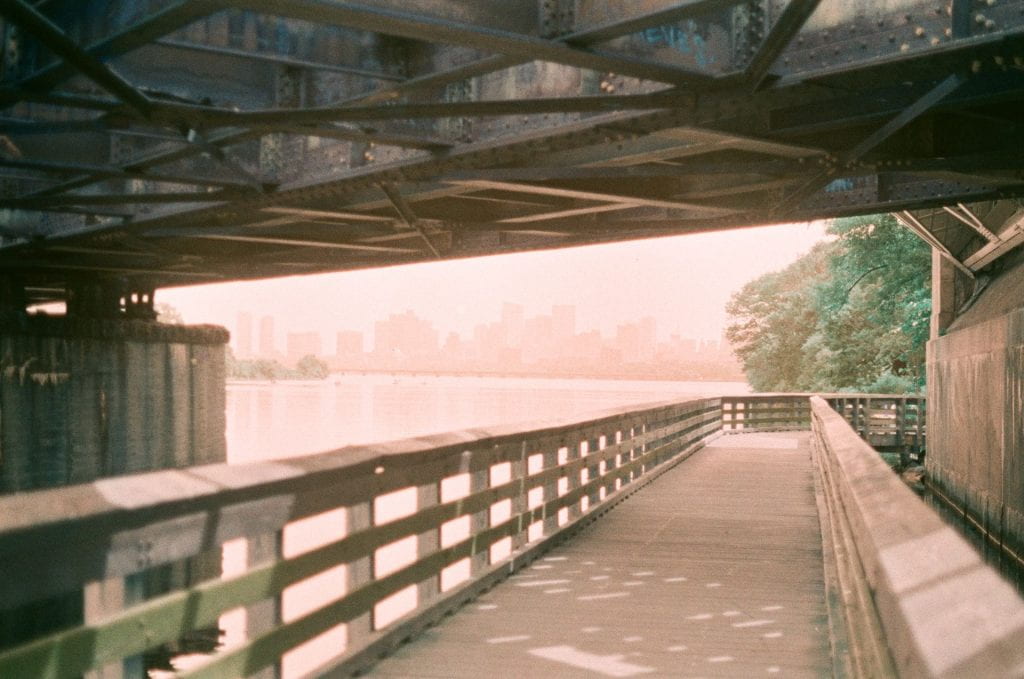 At center, the daytime Boston skyline appears in a pink dreamy haze, faintly reflecting on the calm Charles River. The scene is framed above by the rivets and beams of the BU bridge's underbelly, and framed below by the wooden bridge of the Dr Paul Dudley White Bike Path.