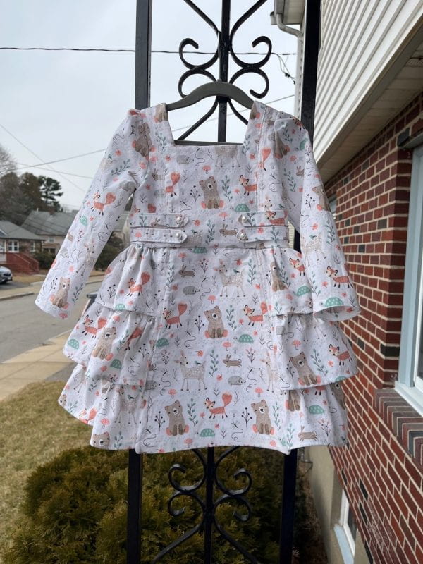A white child's long sleeved dress. The fabric shows drawings of forest critters like bears, foxes and rabbits.