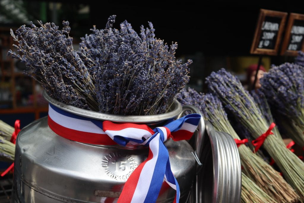 At a market stall, a big bouquet of lavender sprigs is placed inside a tin vessel and smaller lavender bouquets can be seen in the background. Theres is a red ribbon around the small bouquets and a wider red, white, and blue ribbon tied in a bow around the neck of the vessel.