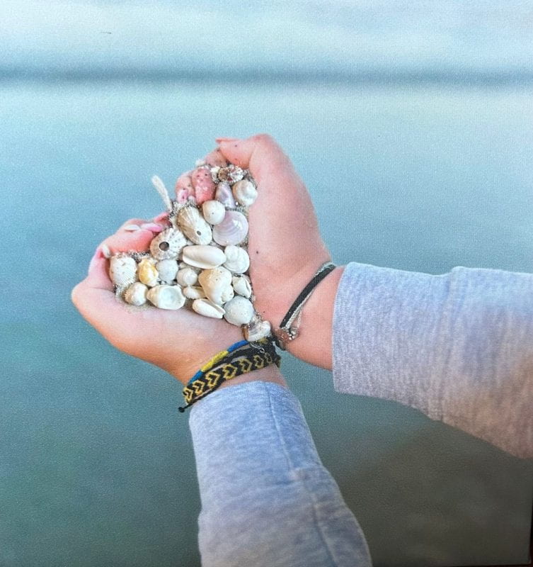 Mixed media of a photo showing the two hands of a person coming together in a heart-shaped handful. Small seashells and sand are glued on top of the photo as the content of the handful.