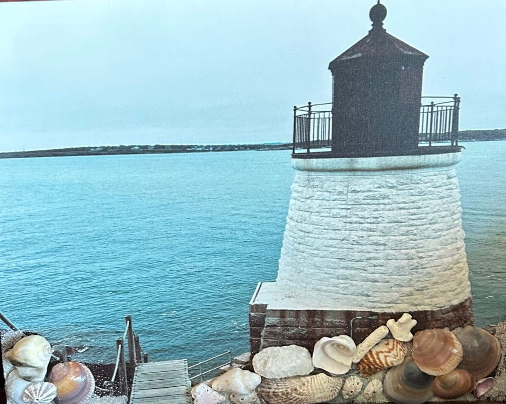 Mixed media with the photo of a lighthouse with white stone walls and a darker top, being framed at the bottom with real-life small seashells.
