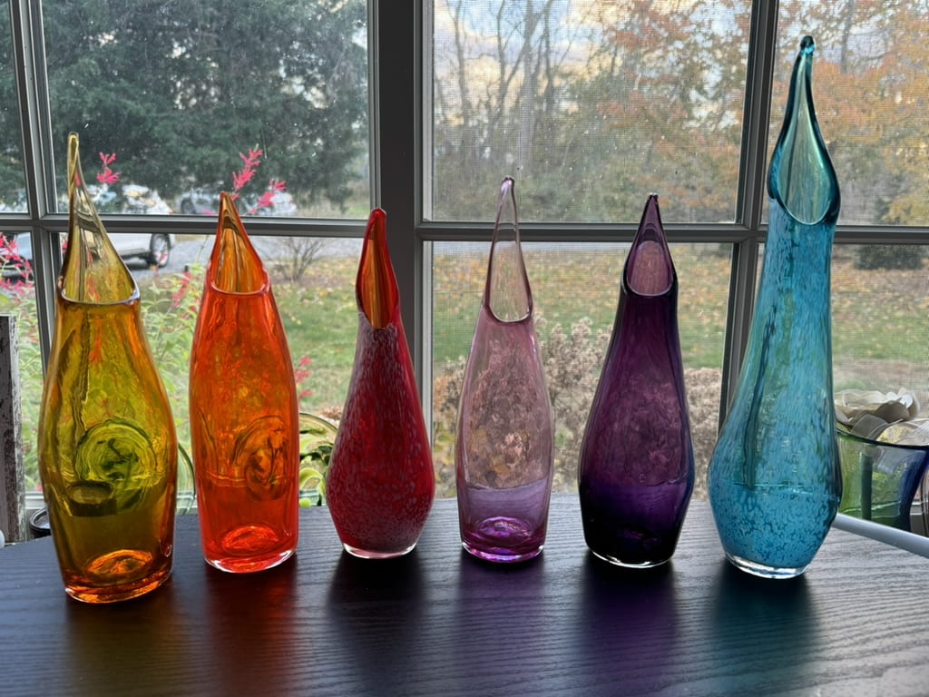 A line of 6 vases - yellow, orange, red, lilac, purple and blue, all inspired by pitcher pants have wide bases and fluted tops that resemble lilies.