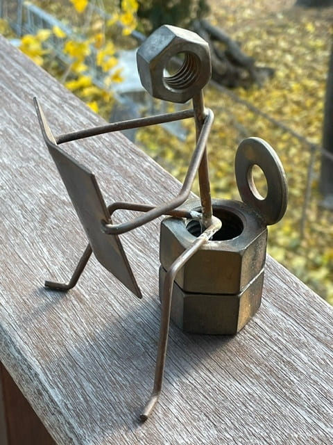 A small metal sculpture rests on an outdoor wooden railing. The sculpture is made of wires, metal nuts, and washers, and depicts a stick figure with a hexnut for a head sitting on a toilet made of two stacked hexnuts, comfortably reading a metal paper.