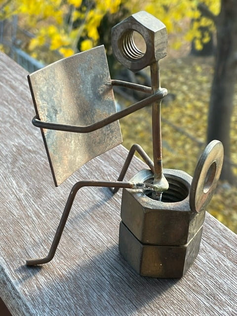 A slightly rotated view of the same small metal sculpture, seen from the "back" of the figure. The sculpture is made of wires, metal nuts, and washers, and depicts a stick figure with a hexnut for a head sitting on a toilet made of two stacked hexnuts, comfortably reading a metal paper.