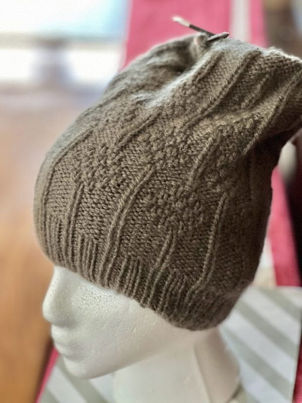 A brown knitted winter hat on a mannequin's head. The knitting needles are still attached to the hat at its top.