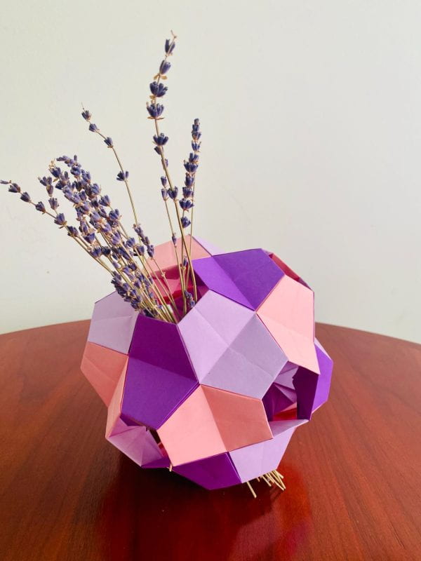 A 3D structure made of small purple, baby pink/orange, and lavender colored paper pieces in the shapes of trapezoids and hexagons. There are numerous holes in the structure. It is used similarly to a vase, with a small bouquet of lavender sprigs going through two of its holes like a tilted axis of the structure.