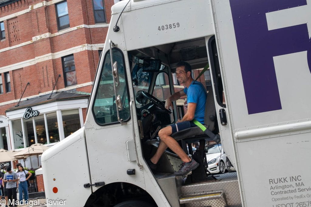 A FedEx driver on Newbury Street taking a break at his wheel, smiling and looking off to the left of the viewer. The photograph is taken at a slight angle.