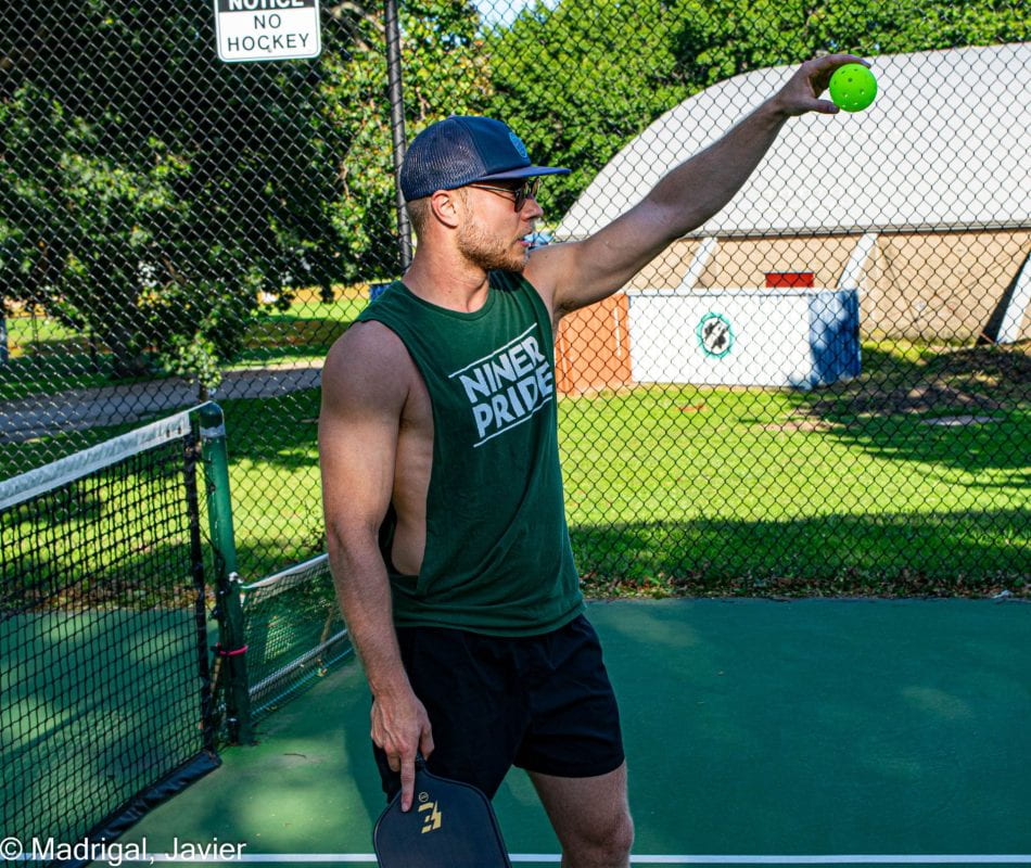 A photograph of a pickleball player readying his shot. He wears a blue hat, dark shorts, and a green tank top reading NINER PRIDE; he holds the green ball high and is addressing someone to the right of the frame.