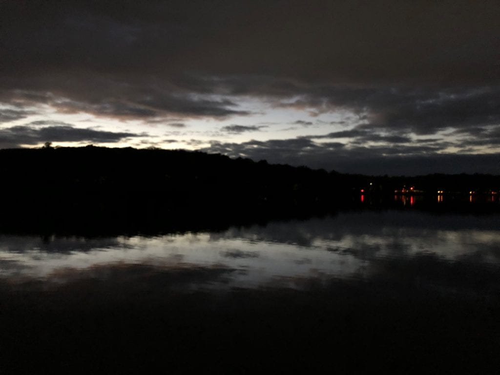 Horizontal image with four layers. The dark clouds at sunset have some grey breaks at the top, with a black layer of hilltop just below, then pond with reflection of the clouds, and finally dark waters on the bottom of the frame. Red and white lights dot the right half of one layer, where car headlights shine in the distance, across the pond.