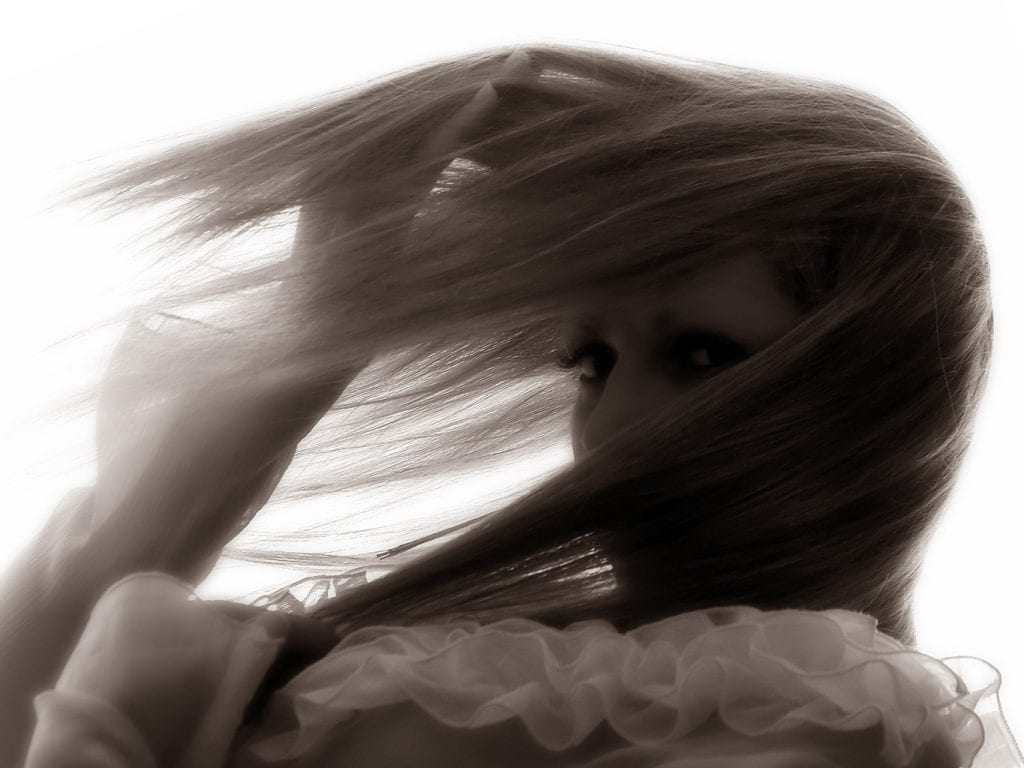 Close-up of a woman in profile who is gazing the camera through her long eyelashes. The wind is coming from her back as seen by the movement of her long hair which covers most of her face. With her two hands she is putting strands of her hair away so that she can keep eye-contact with the camera. The photo is in dark sepia shade.