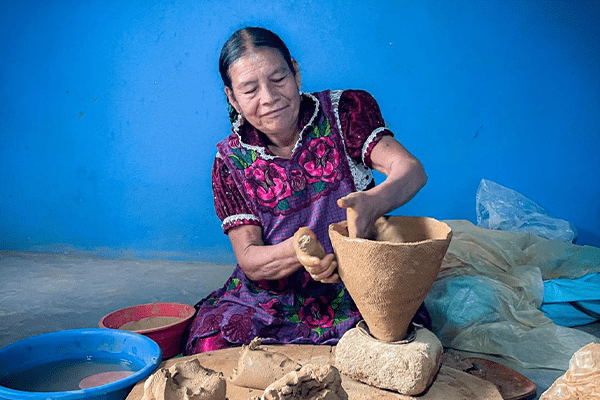 A woman ceramicist looks at her work in the making with a satisfied expression. She is seated down in front of an azure blue wall, wearing a purple dress with magenta flowers.